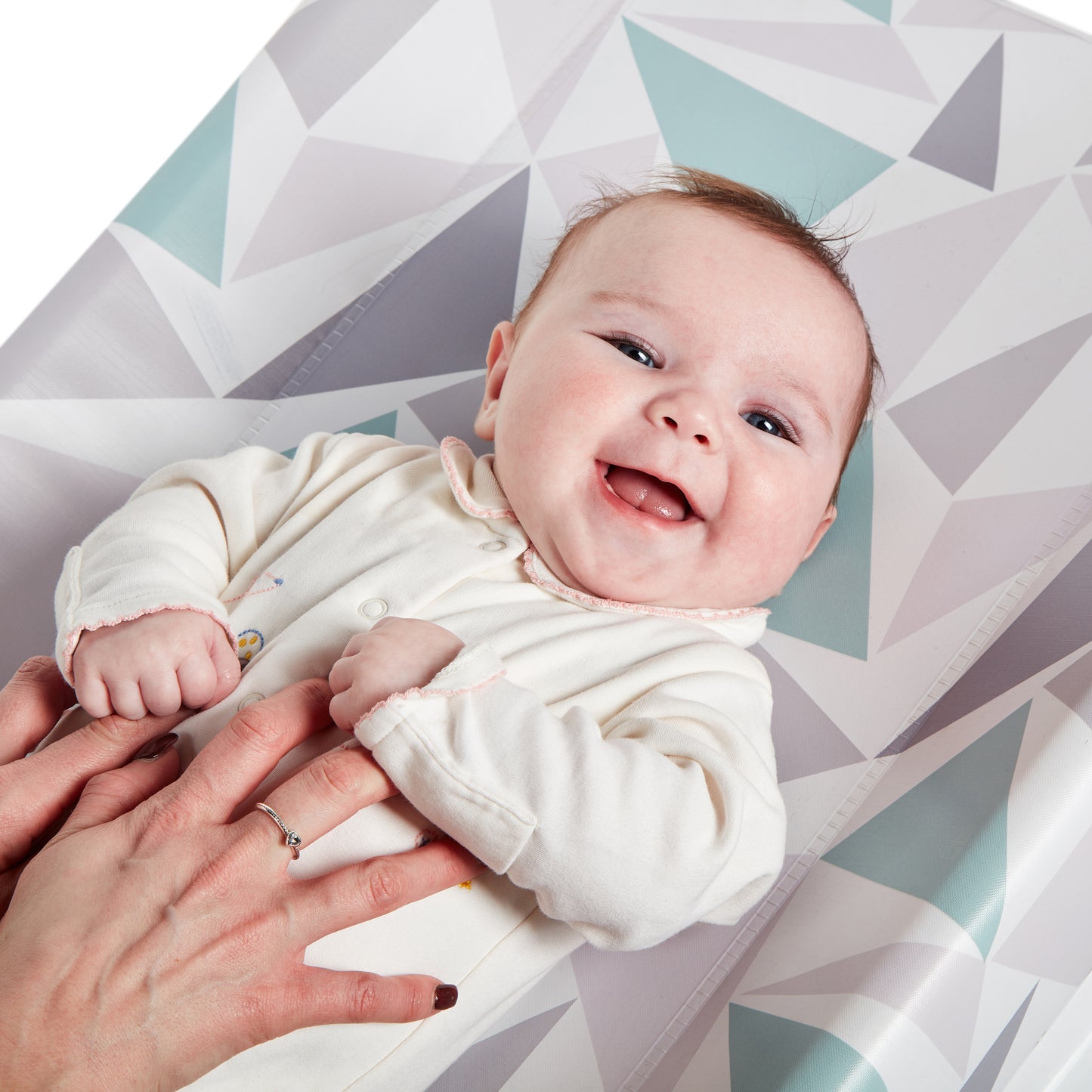 Baby in a white outfit lying on a geometric-patterned mat with a person's hand holding the baby's foot.