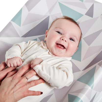Baby in a white outfit lying on a geometric-patterned mat with a person's hand holding the baby's foot.