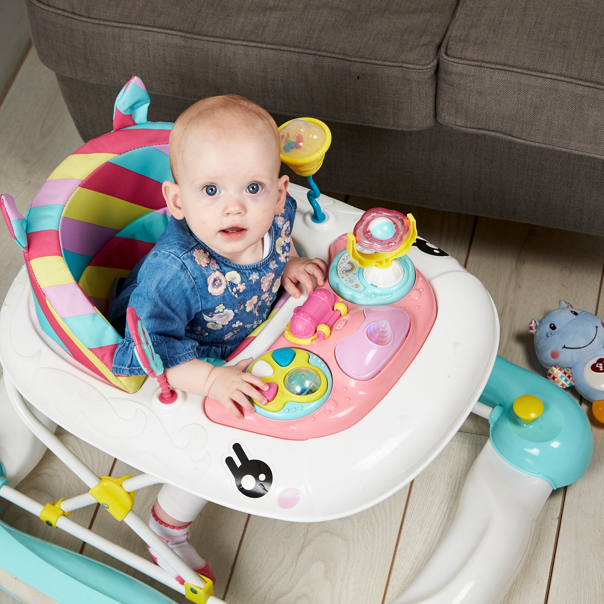 Baby in a colorful activity center on a wooden floor with a grey couch in the background.