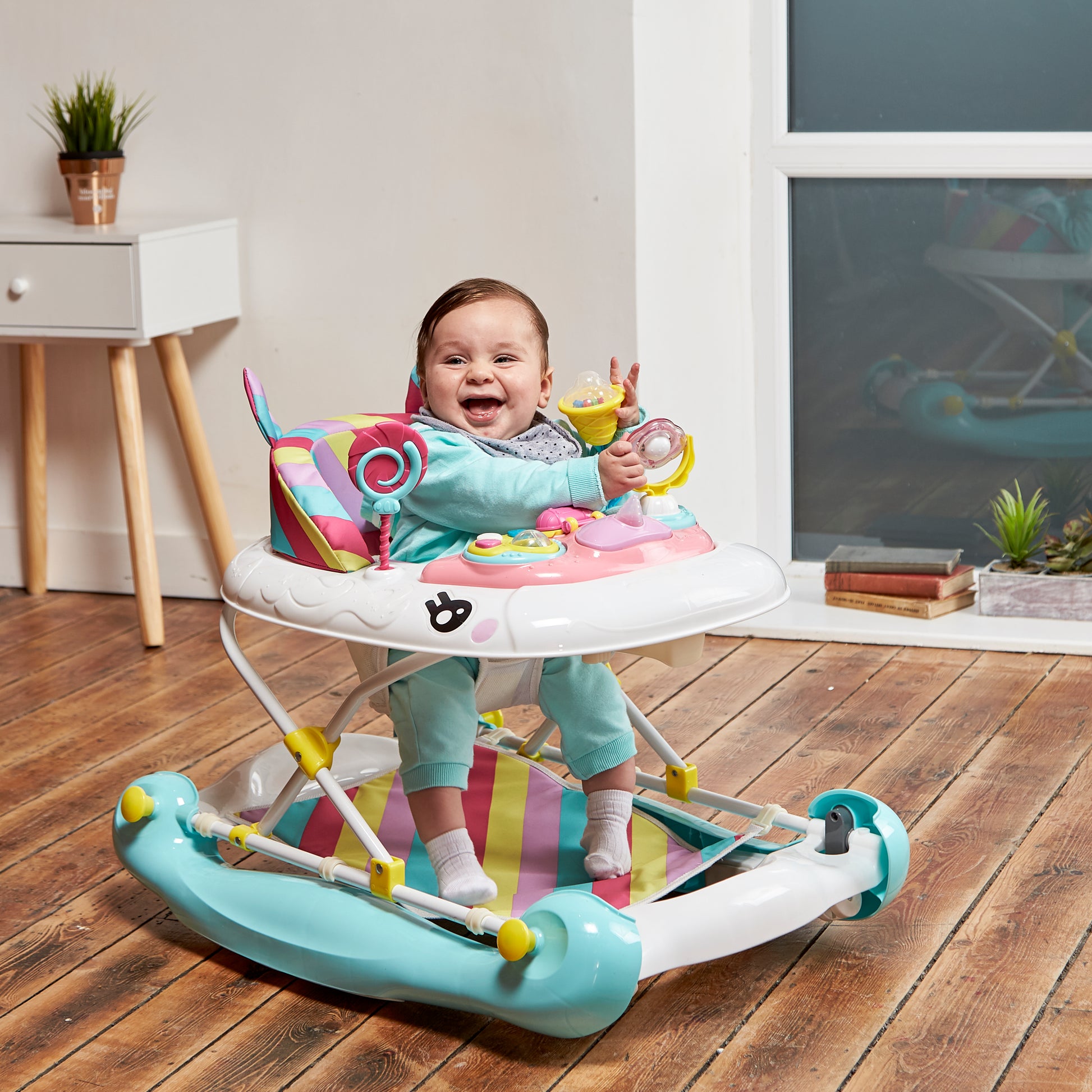 Baby in a colorful walker on a wooden floor with a white wall and window in the background.