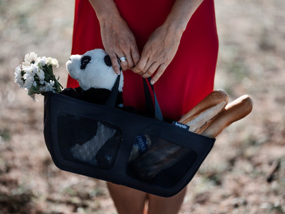 Person holding a black bag with a panda toy, flowers, and bread in a natural setting.
