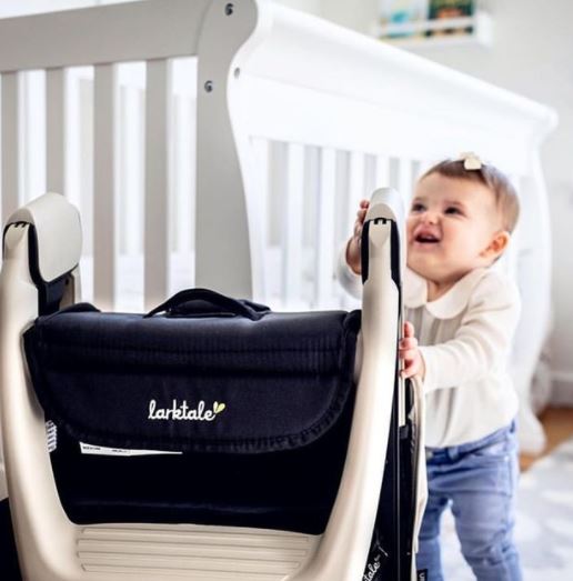 Baby standing next to a high chair with a visible brand logo, in a crib setting.